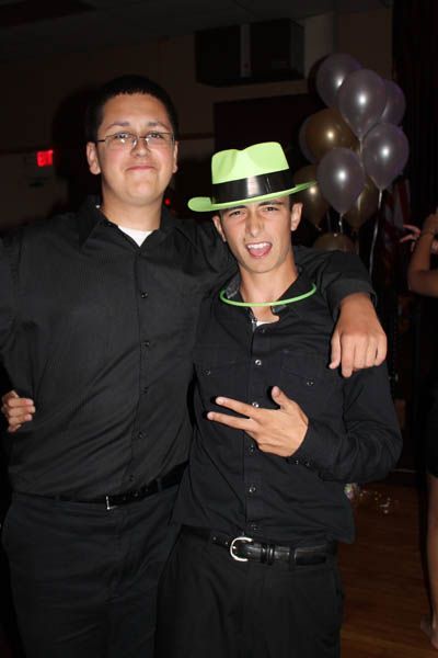 Two young men at an event, one with a neon green hat and gesture. They wear black shirts and pose by balloons.