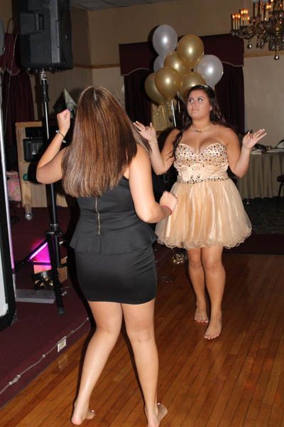 Two barefoot women dancing at a party; one in a black dress, the other in a gold dress. Balloons in background.
