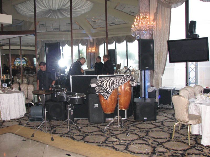 A band setup in a ballroom. Congas, speakers, a DJ setup, and a TV are visible. Three men in black shirts.