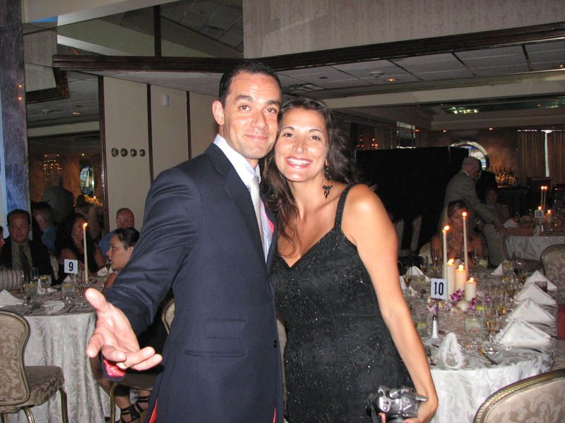 Couple smiling and embracing at a formal event, man in suit, woman in black dress, seated guests in background.