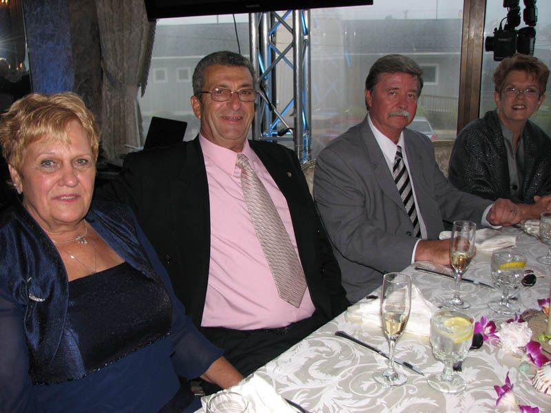 Four people seated at a table, smiling. Men in suits, women with dresses. Drinks and flower centerpieces present.