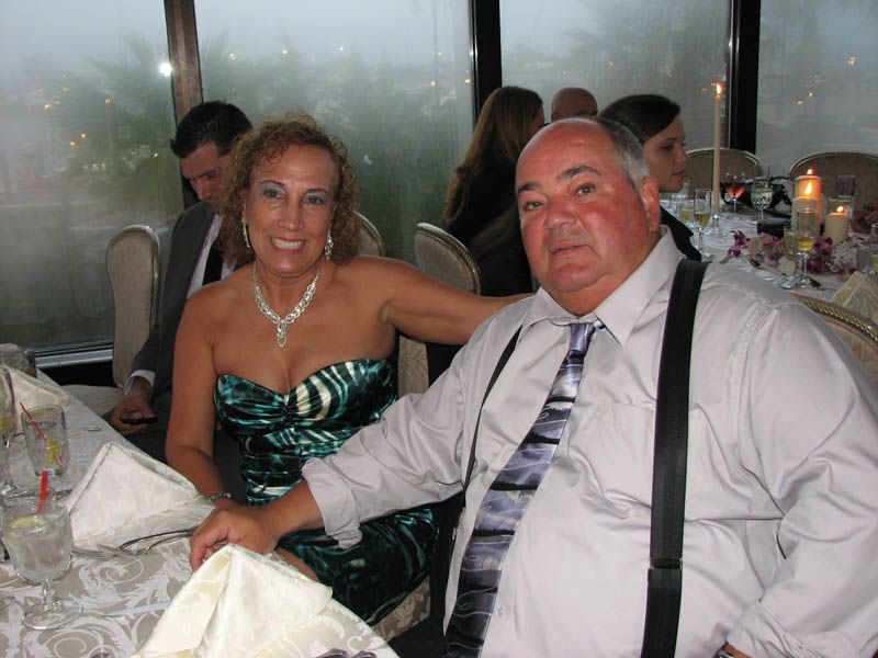 Woman in emerald dress and man in a shirt with suspenders sit at a table. Formal setting with others.