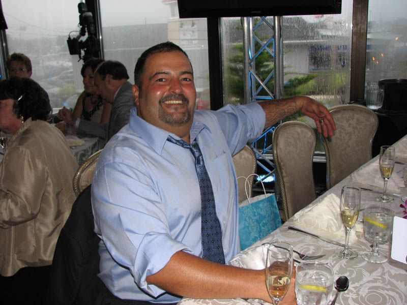 Man at a formal dinner, smiling, leaning on table. Light blue shirt, tie, and champagne glasses.