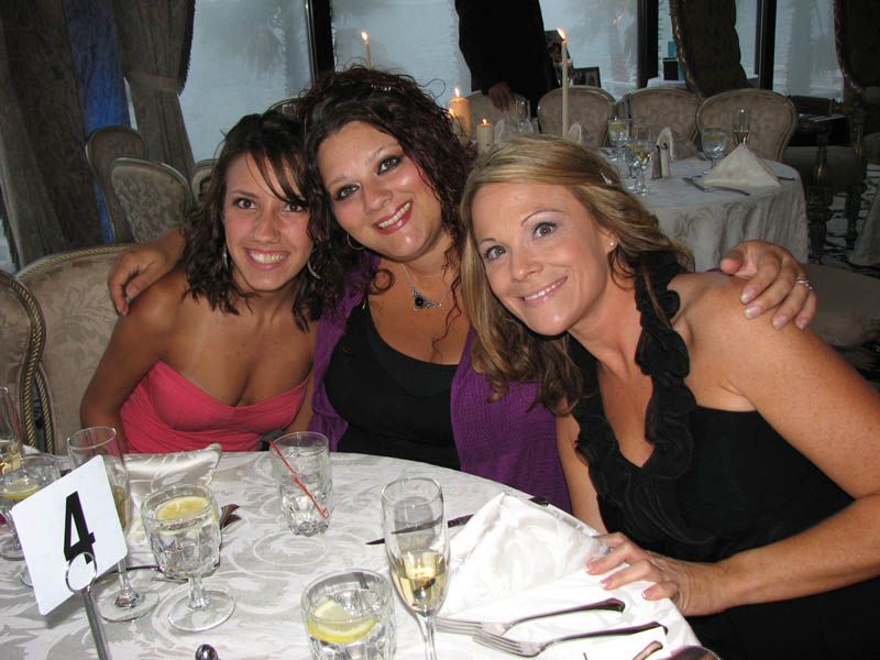 Three women smile at a table at an event. The woman in the center has her arms around the other two.