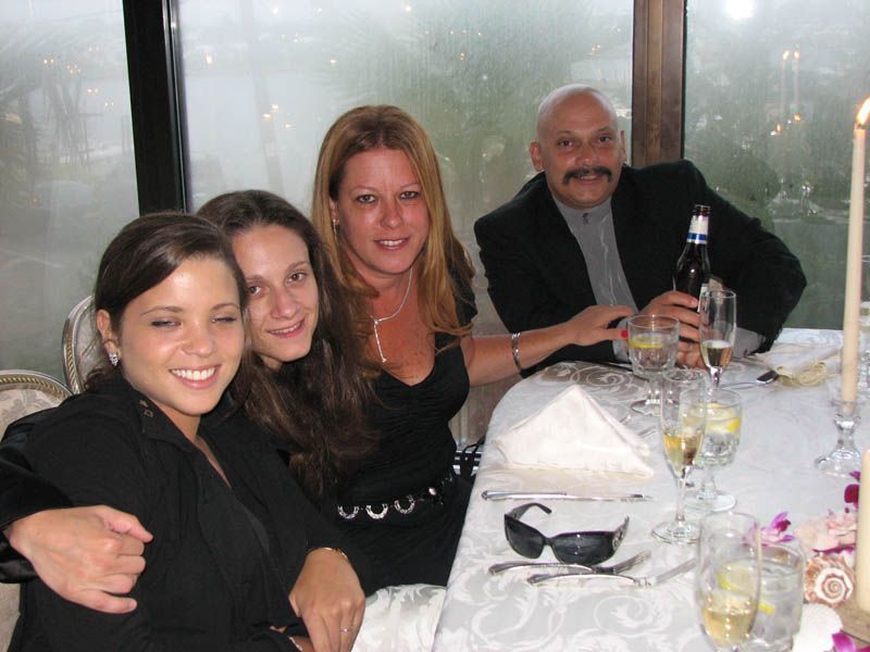 Four people smiling at a table set for a meal. One holds a beer bottle, the others are close together.
