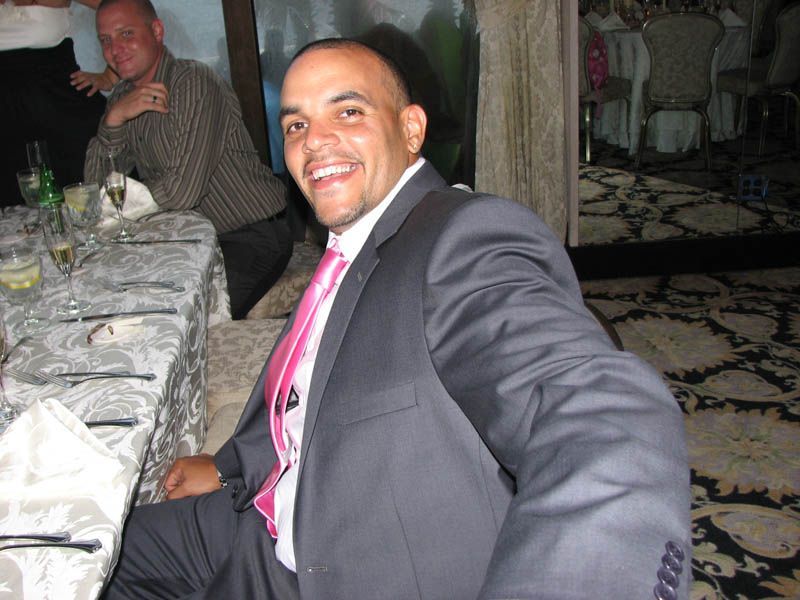 Man in a suit smiles, pink tie, seated at a banquet table. A man in a striped shirt leans in the background.