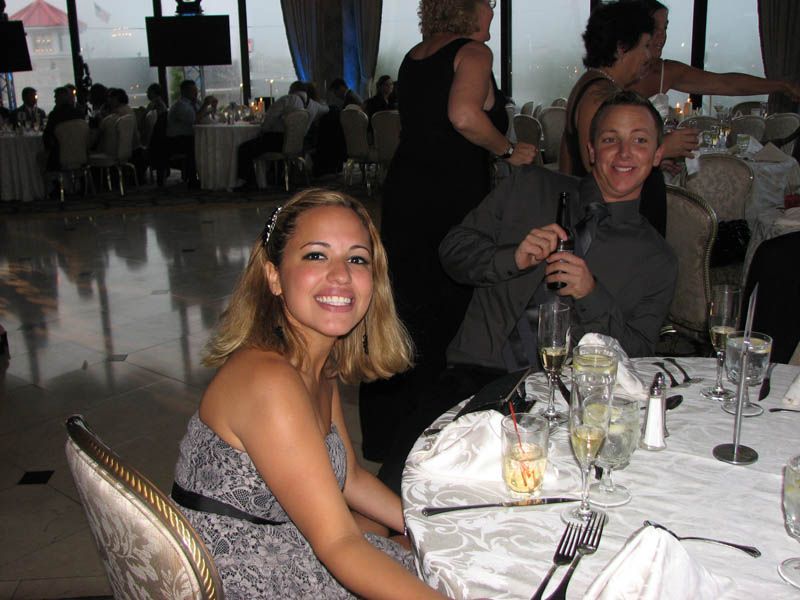 Woman in a formal dress smiles at a table; man smiles and holds a drink. Party setting with tables and guests.