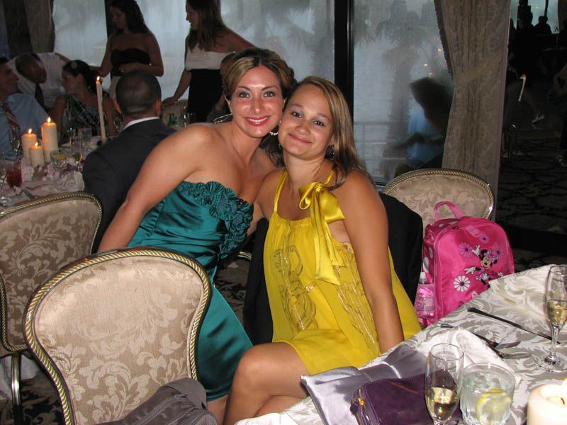Two women smiling at a table, one in a teal dress, one in a yellow dress. Formal setting.