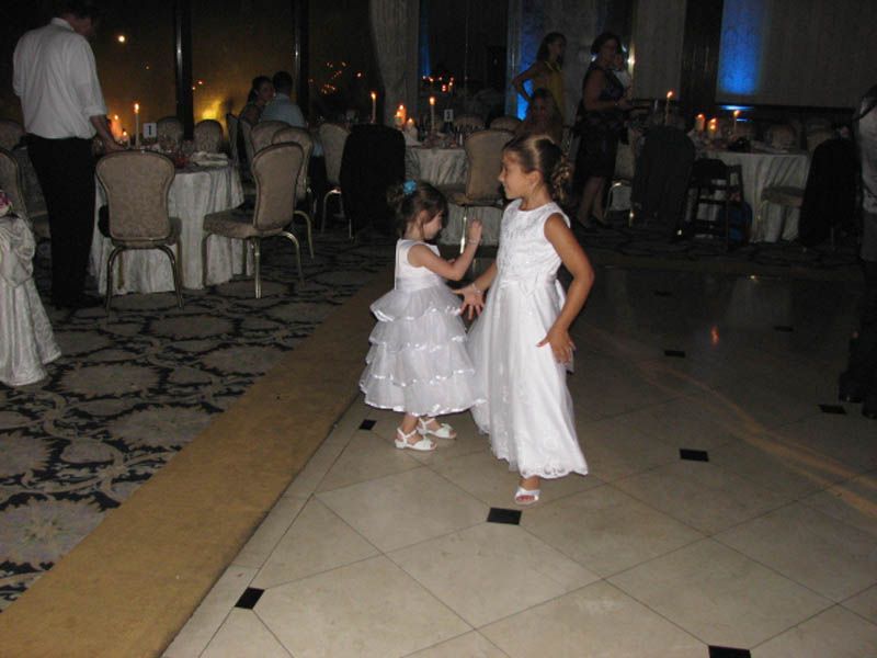 Two young girls in white dresses dancing on a dance floor at an event, tables with guests in the background.