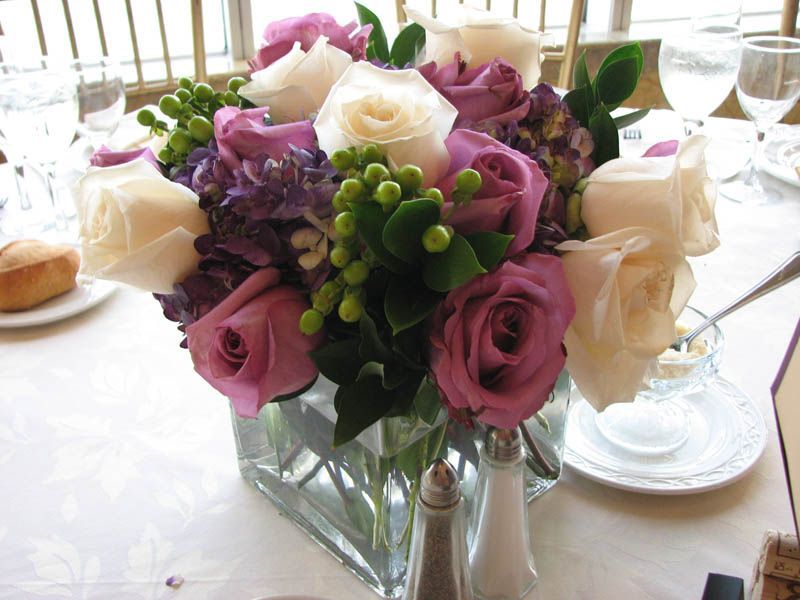 Floral centerpiece with purple and white roses in a glass vase on a white tablecloth.