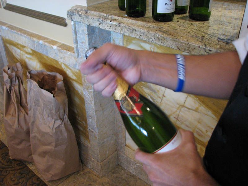 Person opening a green champagne bottle with a white label. Other bottles and paper bags are nearby.