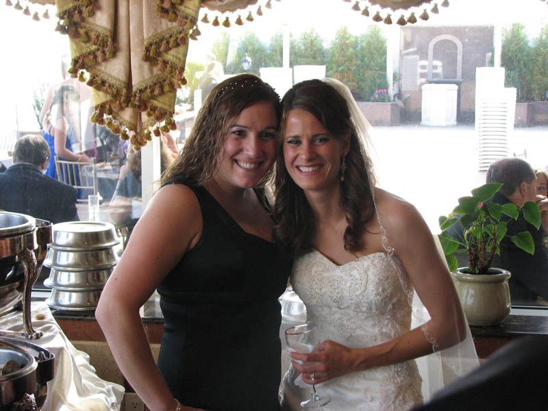 Woman in wedding dress smiles with woman in black dress; wedding reception.
