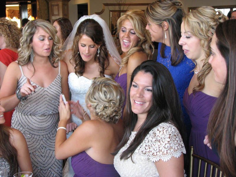 Bride surrounded by bridesmaids; holding cell phone, looking at it, excited expressions, wedding setting.