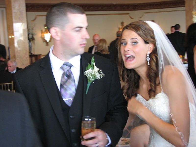 Bride and groom at a wedding, he in a suit with drink, she in a white dress and veil, talking excitedly.