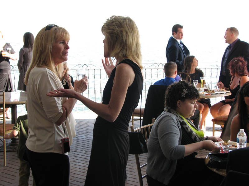 Two women conversing at an event, gesturing with hands. People at tables, sunny outdoor setting.