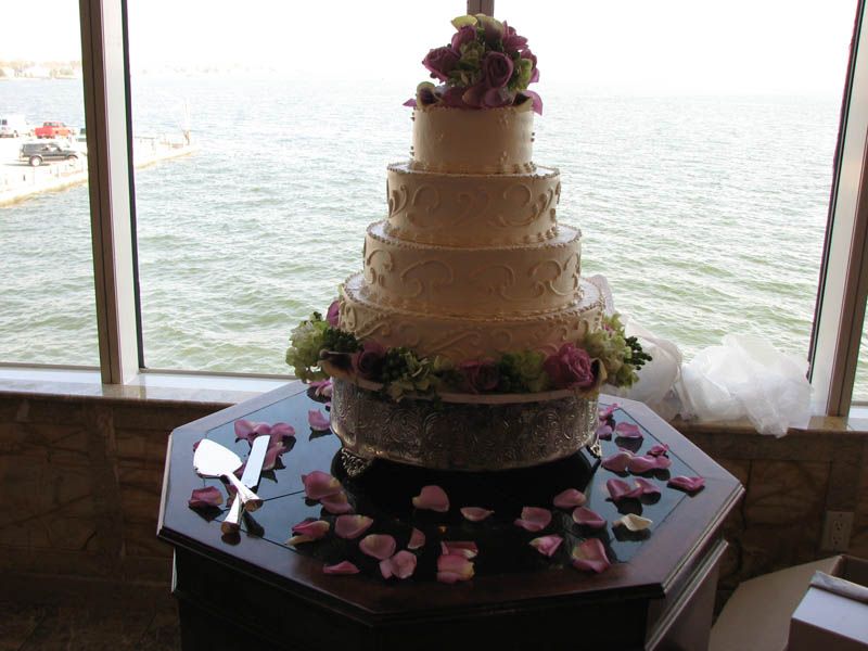 Five-tiered white wedding cake with floral accents, on a decorative stand, next to a window overlooking water.