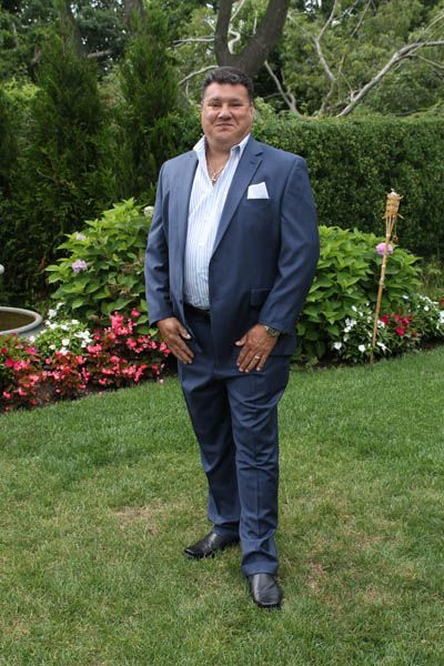 Man in blue suit, open shirt, standing outdoors in a garden, smiling.