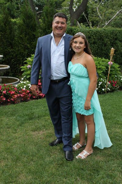Man in a blue suit stands next to a girl in a turquoise dress outdoors, smiling.