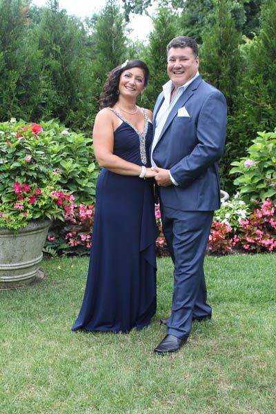Couple holding hands, smiling outdoors. Woman in blue gown, man in suit; lush greenery.