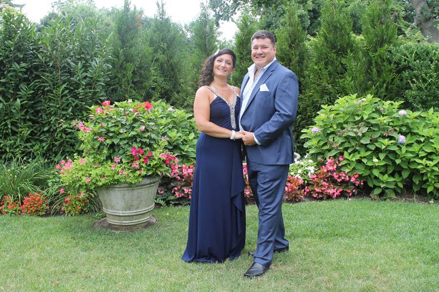 Couple in formal attire pose in a garden, woman in blue dress, man in blue suit.