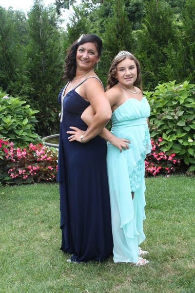 Woman in navy dress and girl in teal dress pose back-to-back outdoors, smiling.