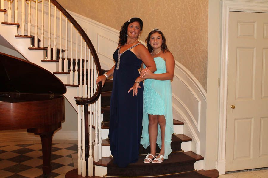 Woman and girl in formal dresses pose on a grand staircase with a piano.