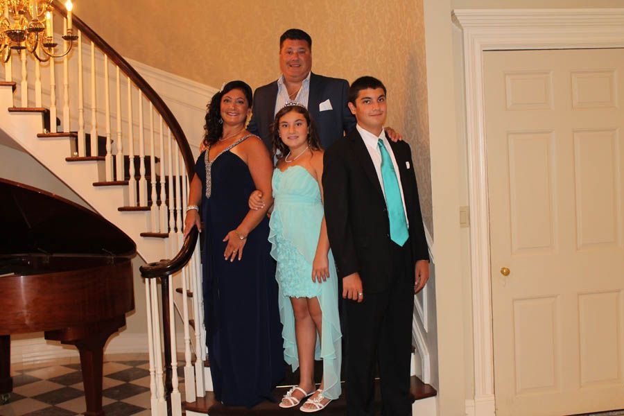 Family poses on a staircase, formal attire. Woman in blue gown, teen in turquoise, man in suit. Piano and door visible.