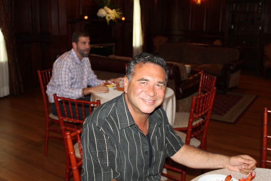 Man smiling at the camera, seated at a table. Another man sits at a nearby table. Formal dining room.