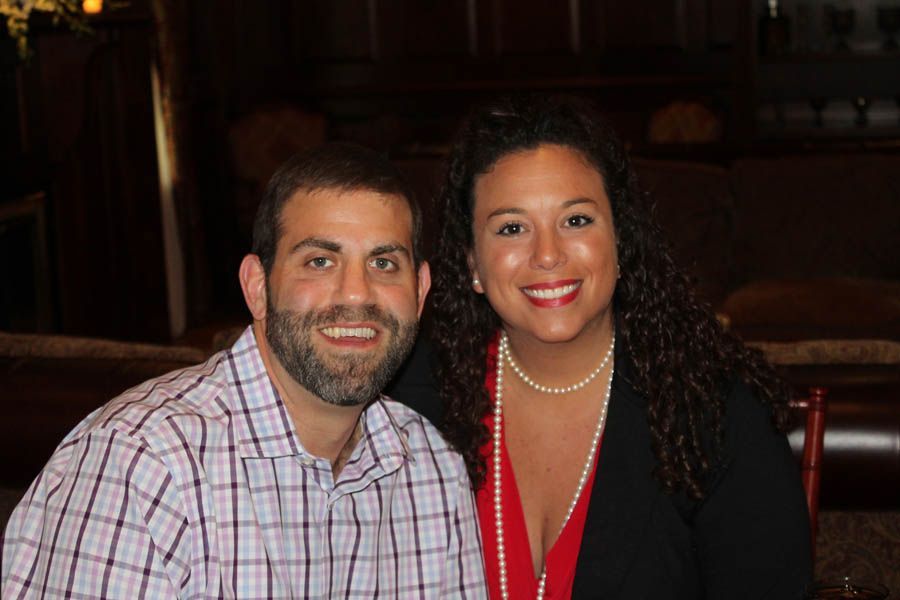 A smiling couple posing indoors; the man has a beard, the woman wears a pearl necklace and a red top.
