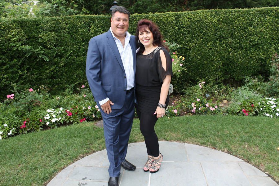 Couple posing outdoors in a garden; man in blue suit, woman in black dress, smiles.