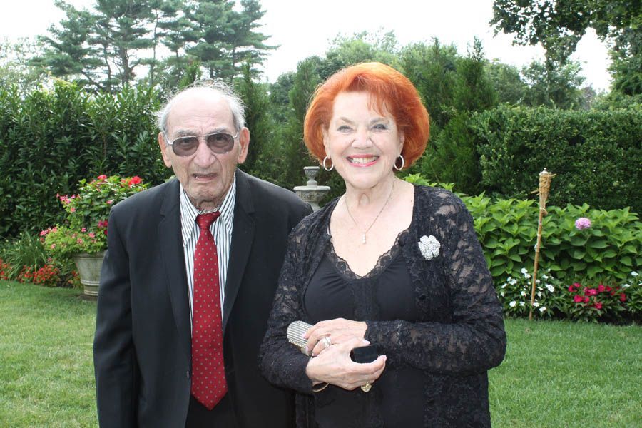 Older man and woman pose outdoors, man in suit, woman in black lace. Lush green garden backdrop.