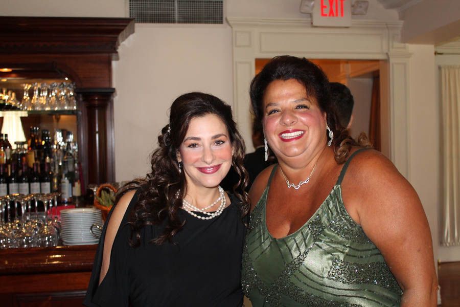 Two smiling women pose together at a formal event, near a bar. One wears a black dress, the other a green beaded gown.