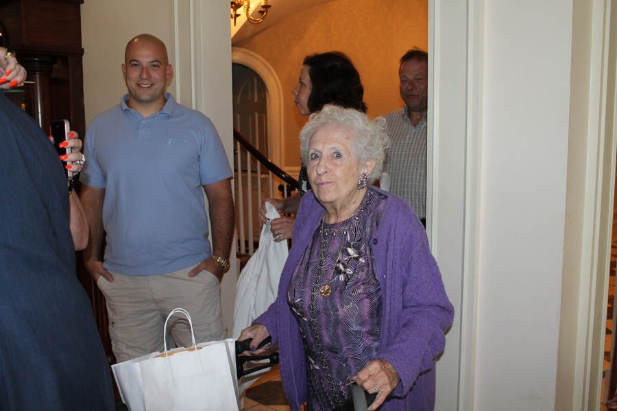 An elderly woman in a purple sweater uses a cane, entering a doorway with people in the background.