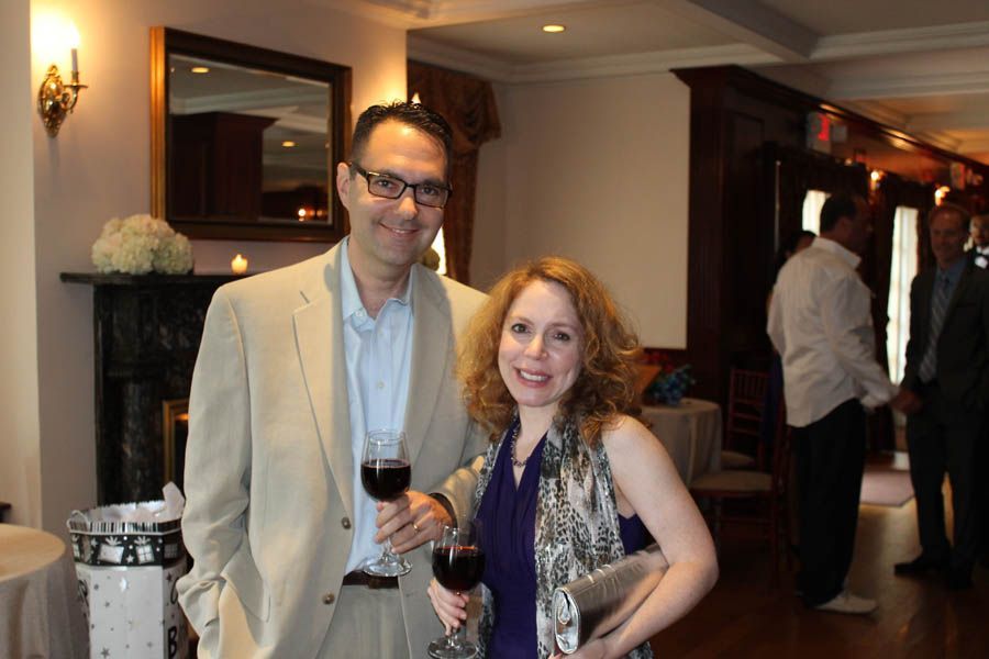 Man and woman at an event, holding wine glasses, smiling. Dark wood interior, other guests in background.