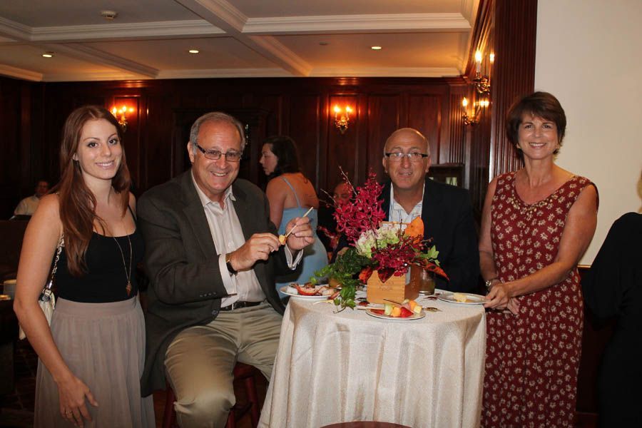 Four people smiling near a table with flowers. Dark wood-paneled room.