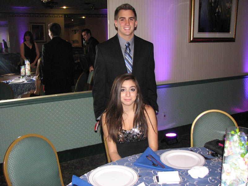 Young man in a suit stands behind a seated woman at a table, both smiling. Formal event setting.