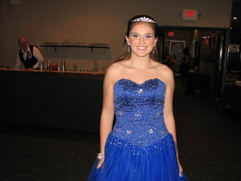 Woman in a blue beaded strapless gown and tiara smiles, standing in a dim room near a bar.