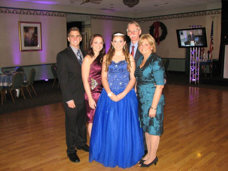 Five formally dressed people pose in a ballroom, a young woman in a blue gown is center.