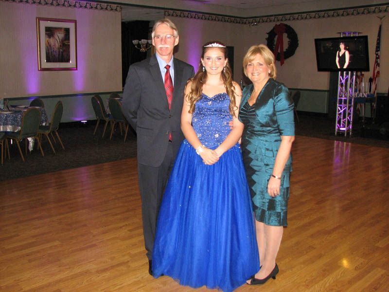 Family poses in a ballroom: girl in blue gown, parents in formal attire, wood floor, decorated room.