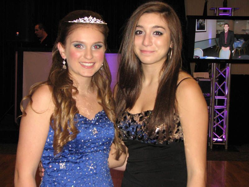 Two young women at a formal event, one in blue gown with tiara, the other in black dress, smiling near a screen.