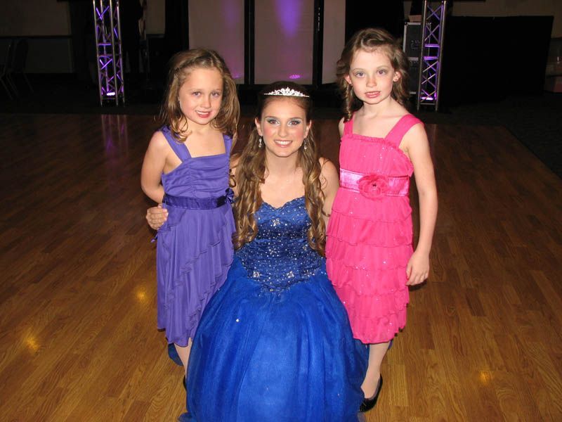 Three girls in formal dresses pose on a wooden dance floor, one in blue, two in purple and pink.
