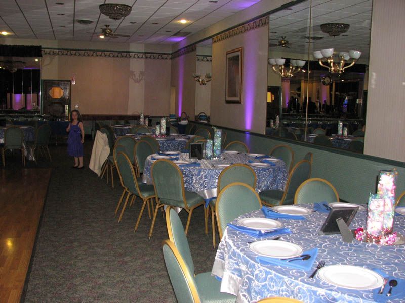 Banquet hall with round tables set for a party, blue tablecloths, green chairs, and a young girl standing.