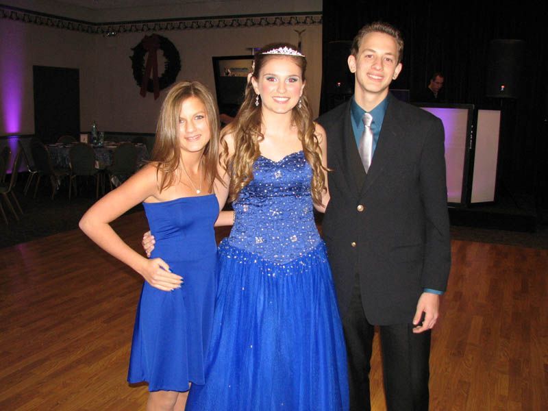 Three teenagers at a formal event, two girls in blue dresses, boy in a suit, posed indoors.