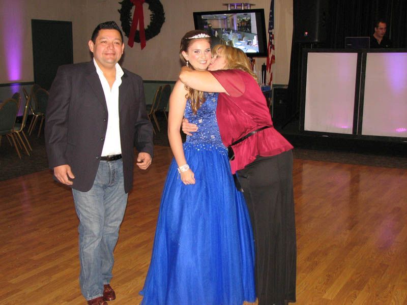A young person in a blue dress with parents, a woman hugging and kissing her, at a formal event.