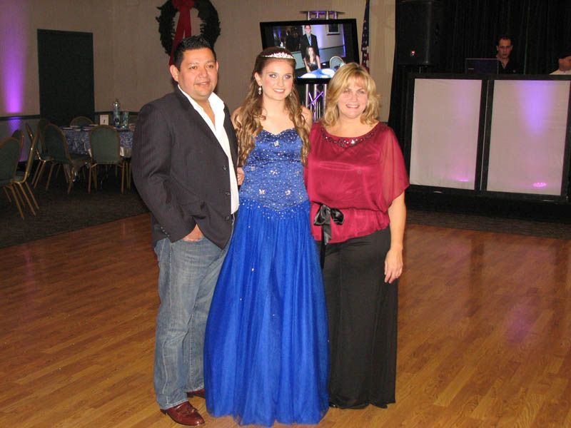 Family poses at an event; woman in a blue gown, man in a blazer, woman in a red top, on a dance floor.