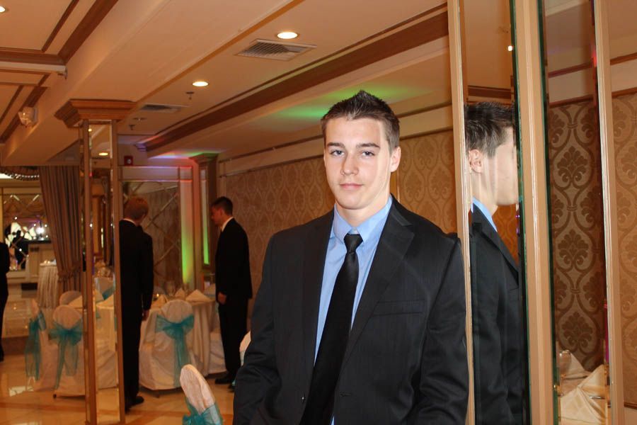 Man in suit standing by a mirror in a decorated room, other people are visible in the background.