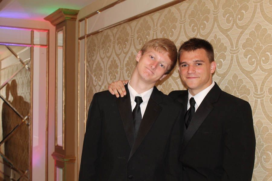 Two men in tuxedos pose, one with arm around the other. Ornate wallpaper backdrop.