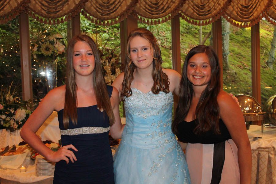 Three young people pose together at an event. One in a light blue gown, others in dark blue and black dresses, smiling.