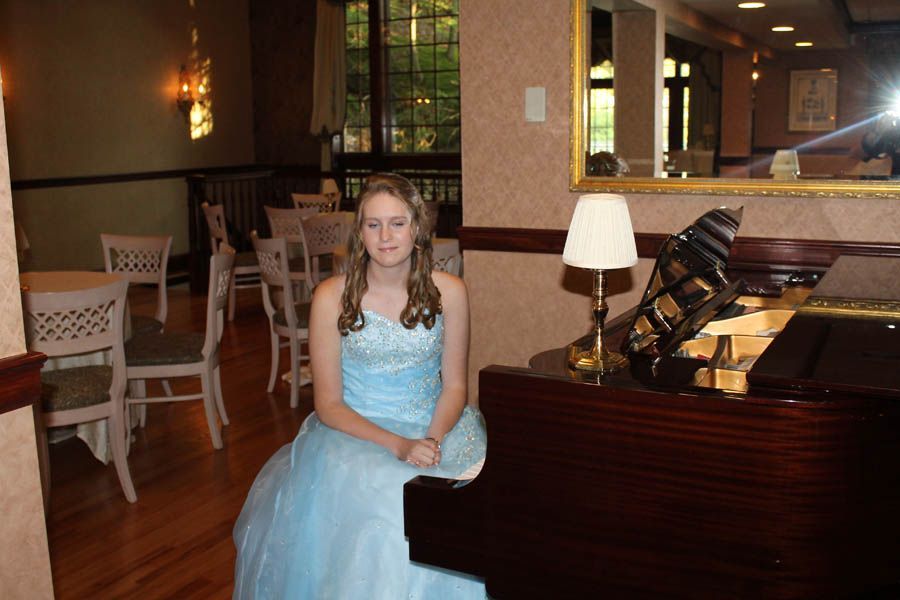 Woman in blue gown seated at a grand piano. She's in a room with chairs and a window.