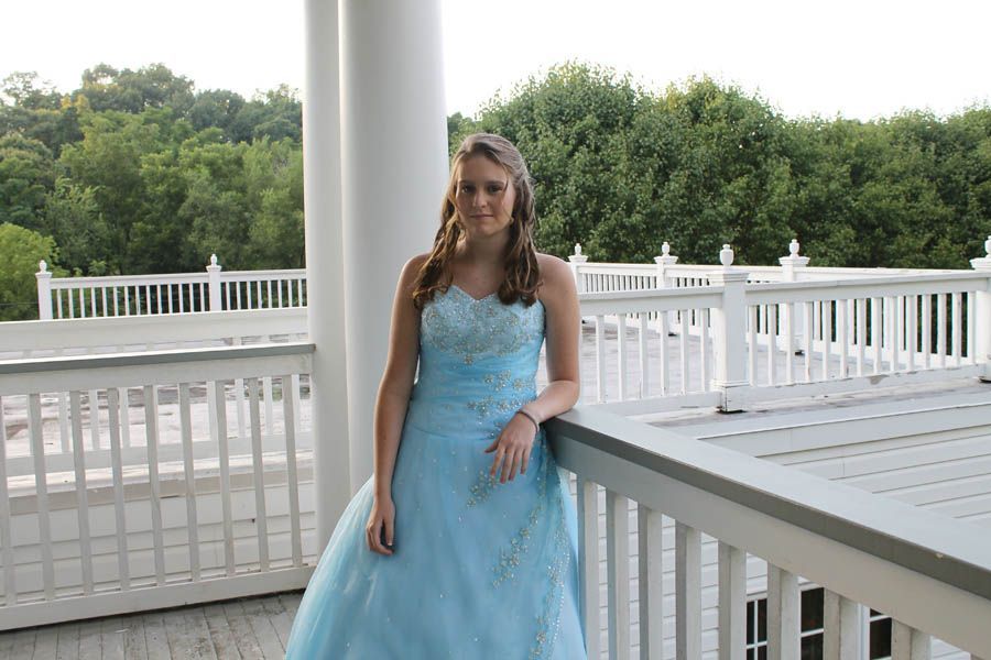 Woman in a blue formal dress on a white porch, trees in the background.